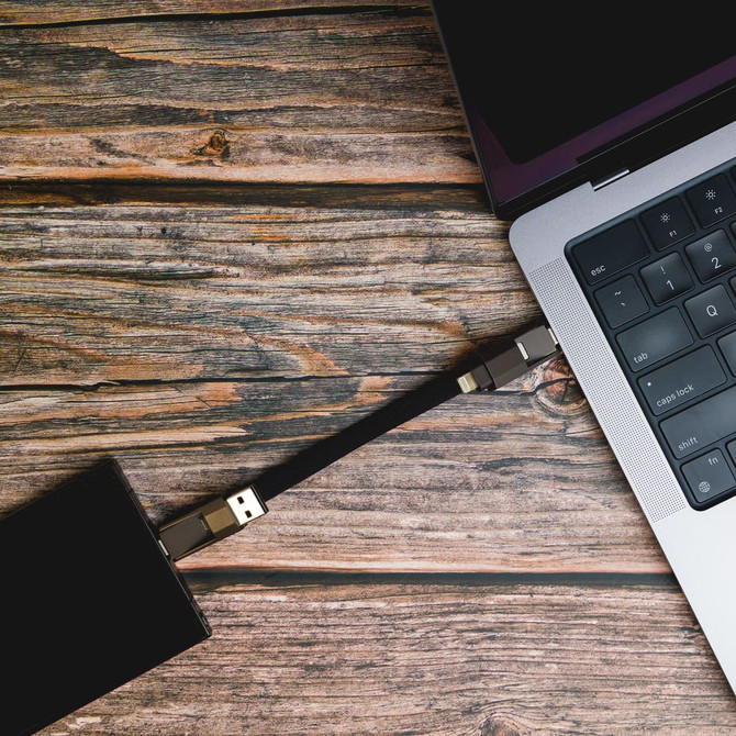 A black charging cable connects a device to a silver laptop on a wooden surface.