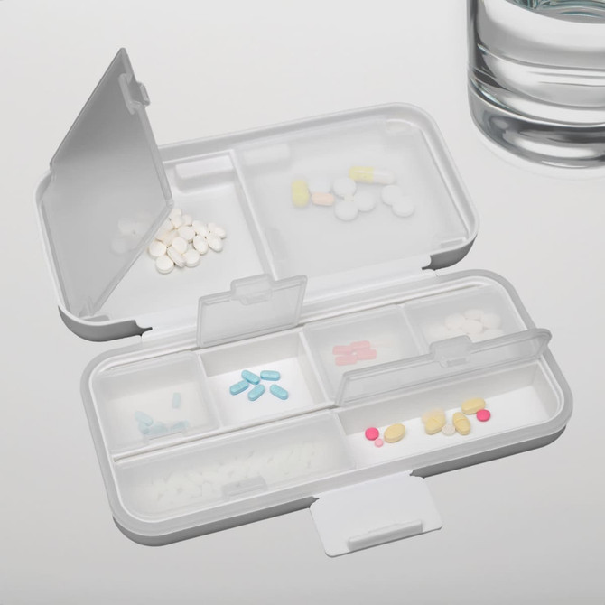 A compact, multi-section pill organiser in white, containing various coloured tablets, next to a glass of water.