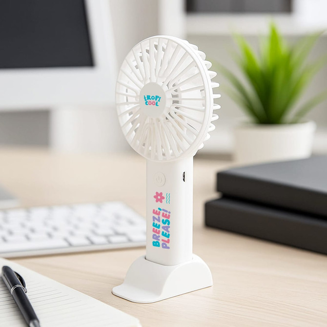 A white handheld fan with a base, featuring a logo and colourful text, placed on a desk near greenery and office supplies.