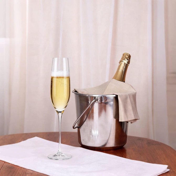A classic champagne flute beside a silver ice bucket holding a champagne bottle, on a wooden table with a white cloth.
