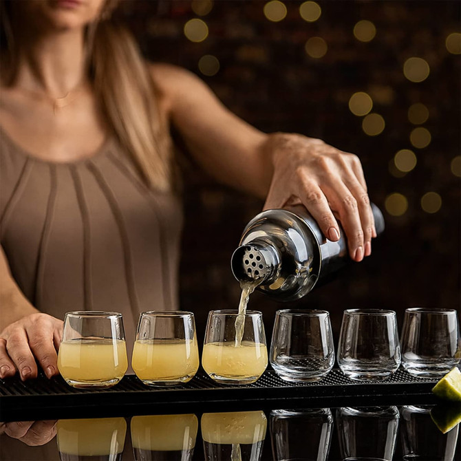 A stainless steel cocktail shaker pouring a light yellow drink into several clear glasses on a bar countertop.