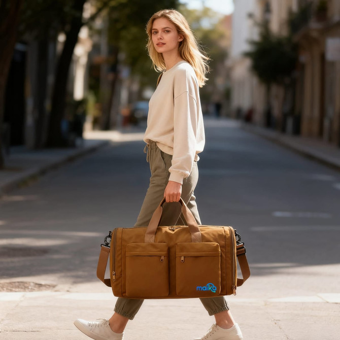 A woman carrying a foldable travel duffle bag in brown while walking on a street. The bag features a logo.