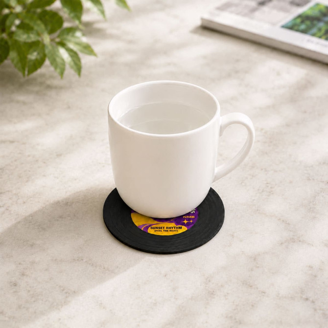A white mug sitting on a black custom record coaster on a marble surface, beside a green plant and a magazine.