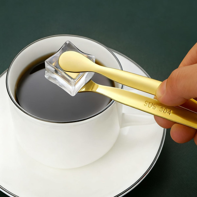 A pair of gold stainless steel ice tongs grasping a clear ice cube above a cup of black coffee.