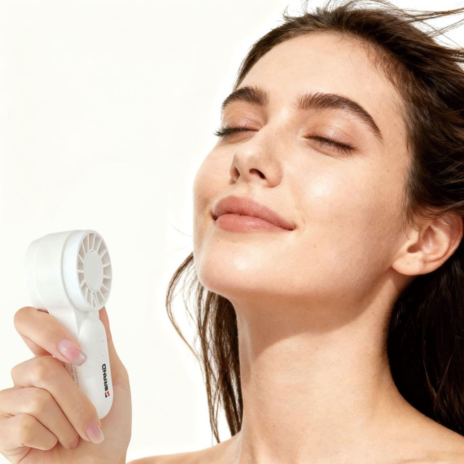 A white portable handheld fan is being held by a woman with a relaxed expression and flowing hair.