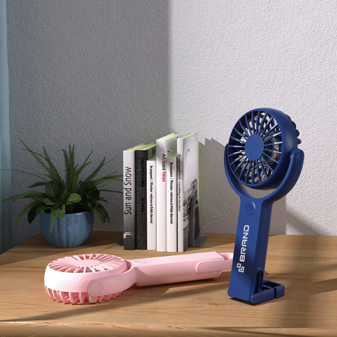 A blue and pink handheld fan with a 180° adjustable angle, placed on a wooden surface near books and a plant.