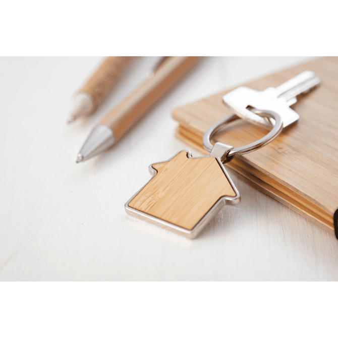 A bamboo keyring in a house shape, featuring a silver keyring attachment, placed next to pens and a wooden surface.