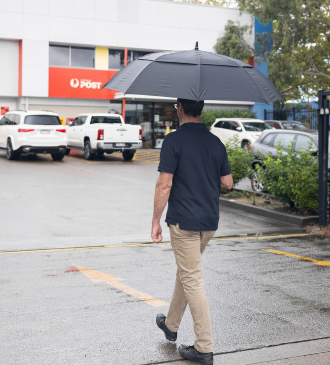 A person walking under a large black golf umbrella, with cars and a post office in the background.