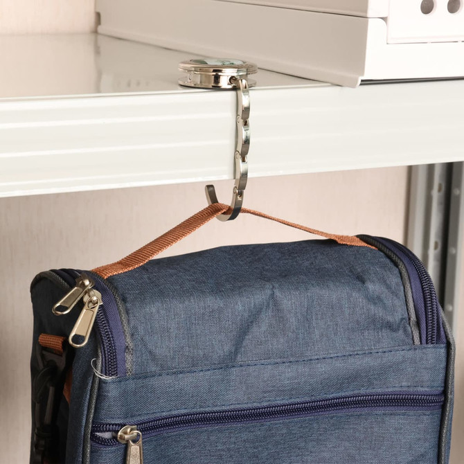 A foldable desk hook in silver holds a blue bag on a shelf. The hook features a mirror design.