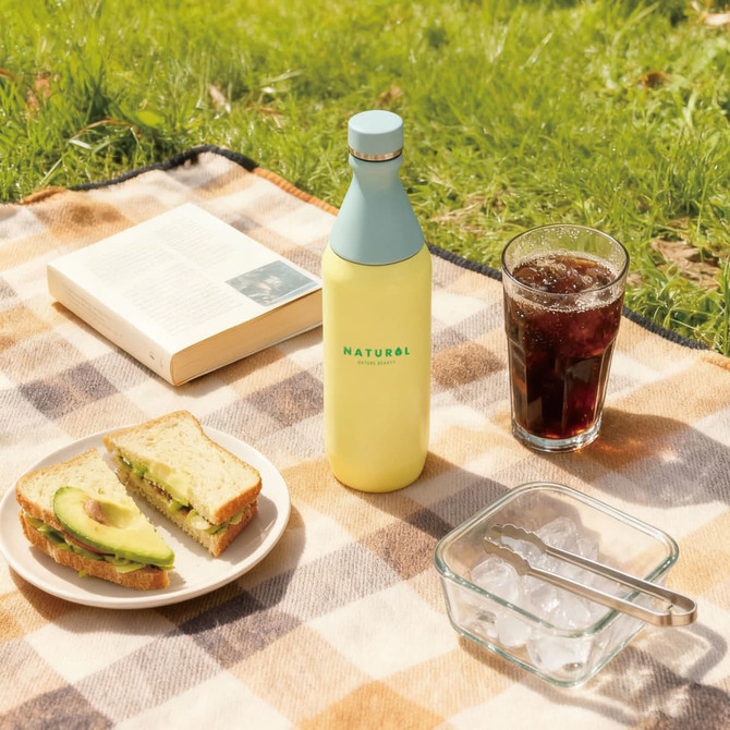 A drink bottle in pale yellow with a light blue cap, set on a picnic blanket with food and a beverage.