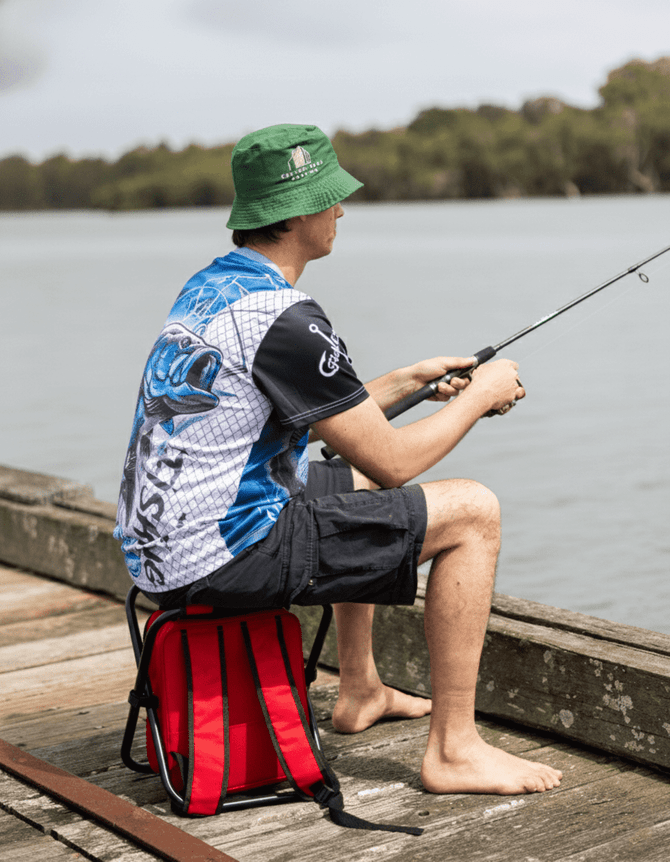A man wearing a blue and white polyester fishing tee and a green bucket hat sits by the water, fishing while seated on a red bag.