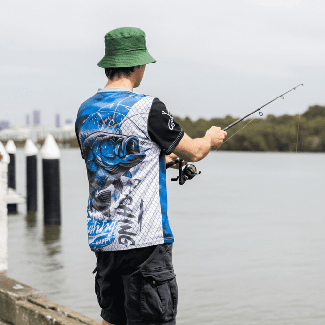 A man in a blue and black fishing tee stands by the water, holding a fishing rod. He wears a green bucket hat.