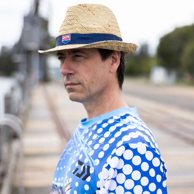A man wearing a straw hat and a blue patterned long sleeve fishing tee stands by a waterway with a serene expression.