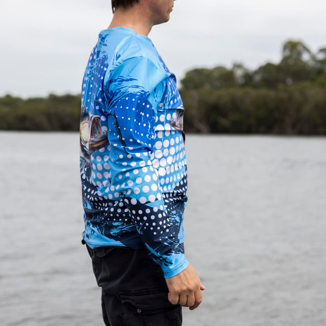 A long sleeve sublimated fishing tee in blue, featuring a fish design, displayed against a waterfront backdrop.
