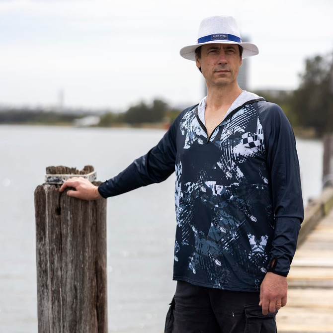 A man stands by the water wearing a navy and white polyester sublimated 1/4 zip fishing hoodie, with a hat and shorts.
