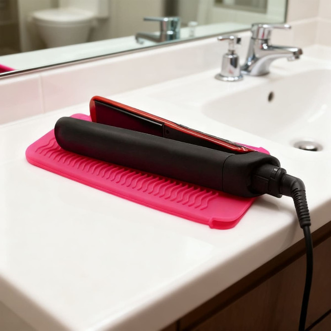 A black hair straightener and a red styling tool rest on a pink silicone mat on a bathroom countertop.