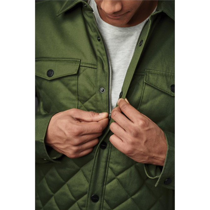 A man in a green quilted jacket adjusts the zipper, revealing a grey top underneath. The jacket features snap buttons and a logo.