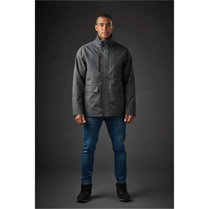 A man wearing a grey Montauk System jacket paired with blue jeans and black shoes, against a dark background.