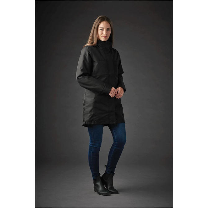 A woman is wearing a long black jacket with a high collar, paired with blue jeans and ankle boots. The background is dark.