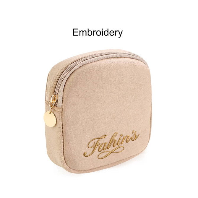A beige mini makeup bag featuring a zip closure and gold embroidery, with a small round charm attached.