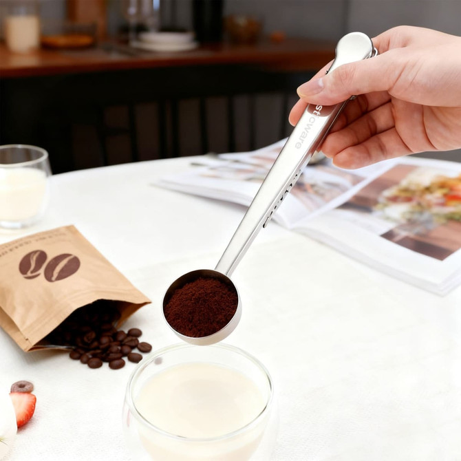 A stainless steel coffee measuring spoon with a clip, holding ground coffee above a glass of milk on a table.
