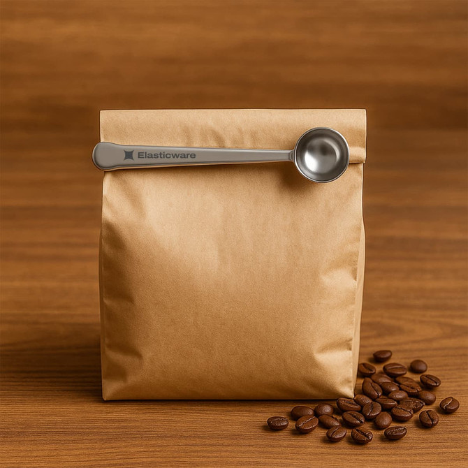 A brown paper bag of coffee with a stainless steel measuring spoon resting on top, surrounded by coffee beans.