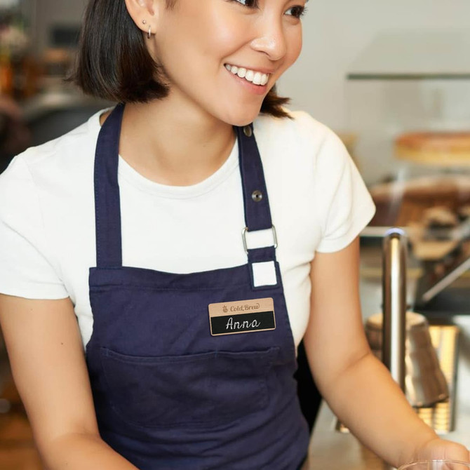 A wooden chalkboard name badge attached to a navy apron worn by a smiling person in a café setting.