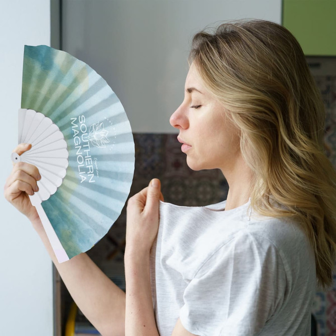 A fabric hand-held fan featuring a gradient design in soft blue and green colours, being held by a woman with blonde hair.