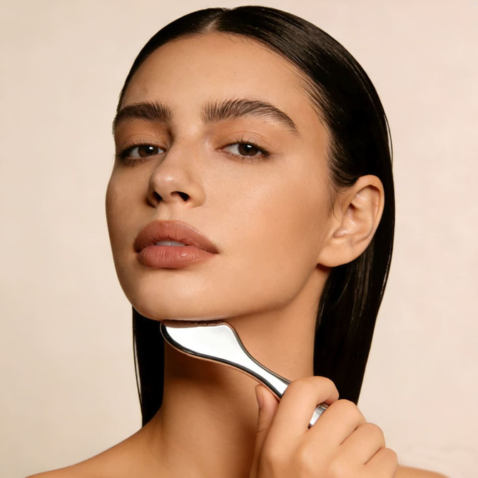 A woman with long, dark hair gently holds a silver facial tool against her jawline, showcasing her natural beauty.