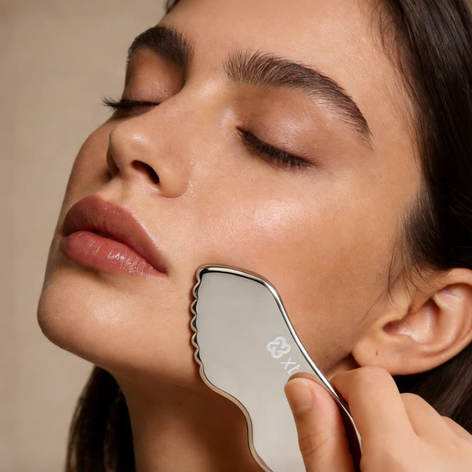 A customised Gua Sha board with a sleek, silver design is being used on a woman's face.