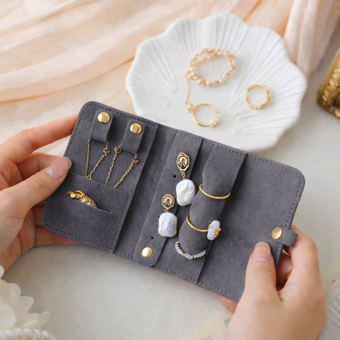 A grey synthetic leather jewellery bag displaying various rings and earrings, held open in two hands.