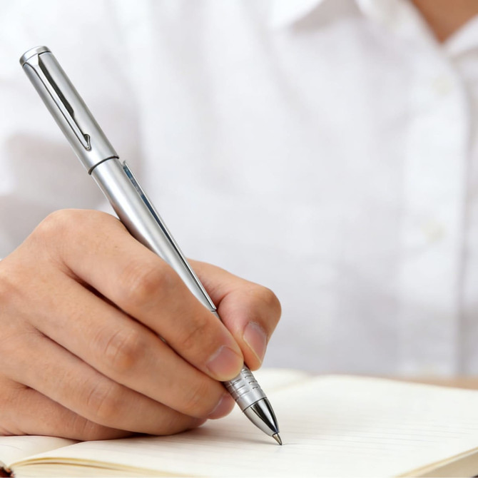 A silver pen held in a hand, poised above a blank notebook on a table.