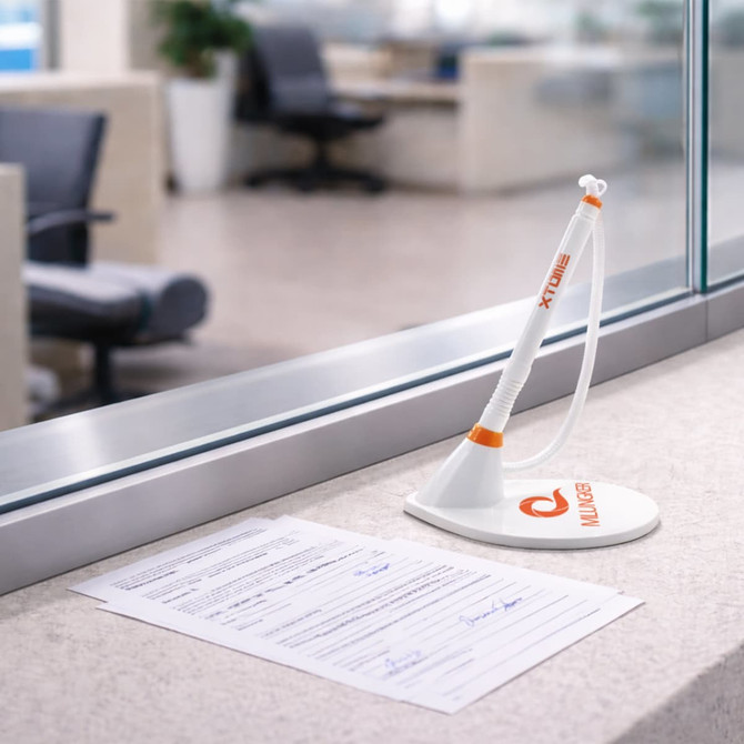 A fixed signature pen in white and orange stands on a countertop beside a document with signatures.