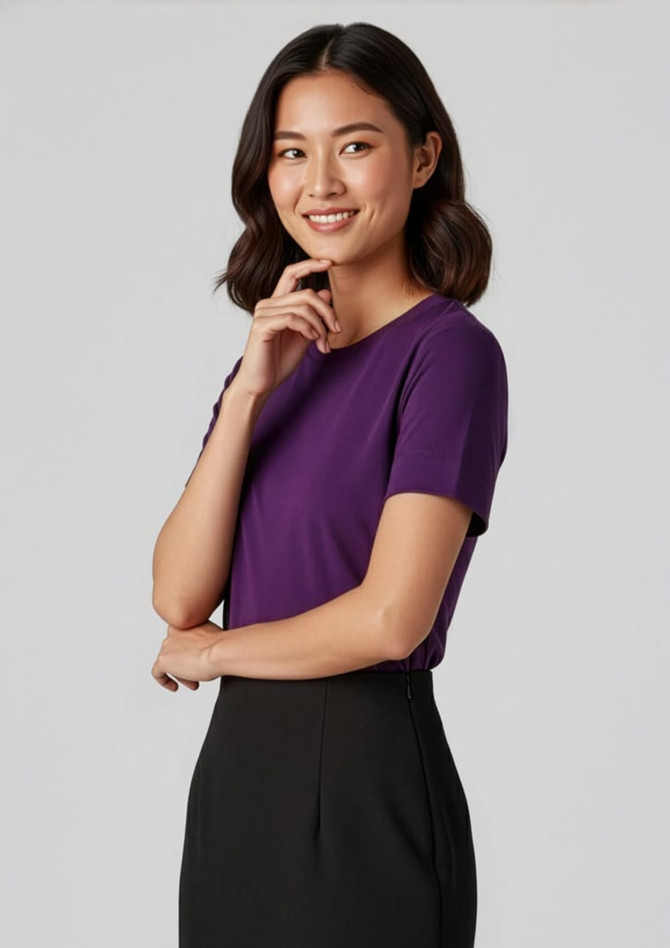 A woman models a purple short sleeve jersey top, paired with a black skirt. The top features a subtle logo.