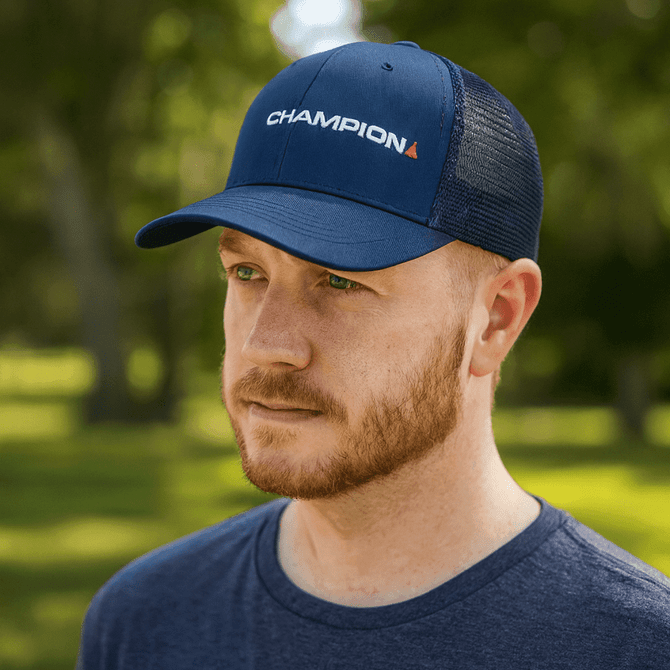 A navy snapback trucker cap with a mesh back and a logo, worn by a man with a beard in a natural setting.