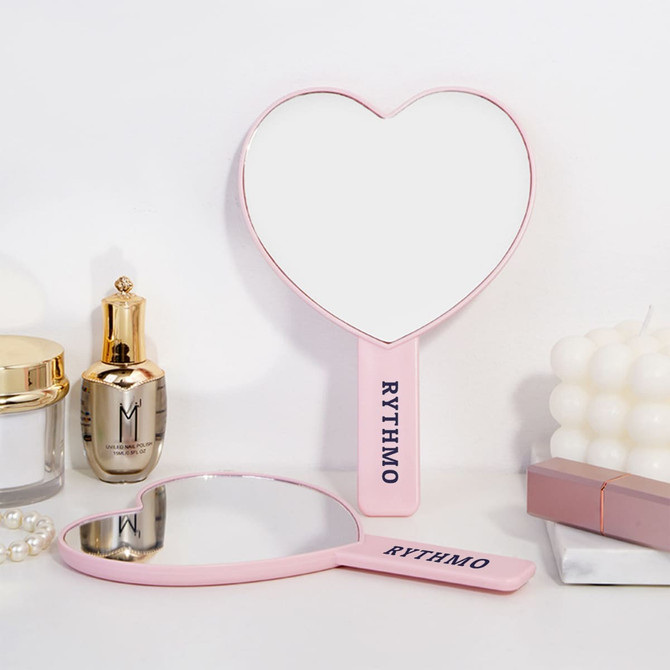 A heart-shaped handheld mirror with a pink handle and a logo, surrounded by beauty products and candles.