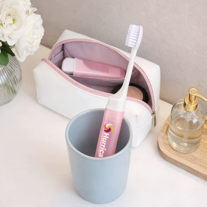 A pink transparent foldable handle toothbrush is placed in a grey cup next to a white and pink cosmetic bag and a glass bottle.