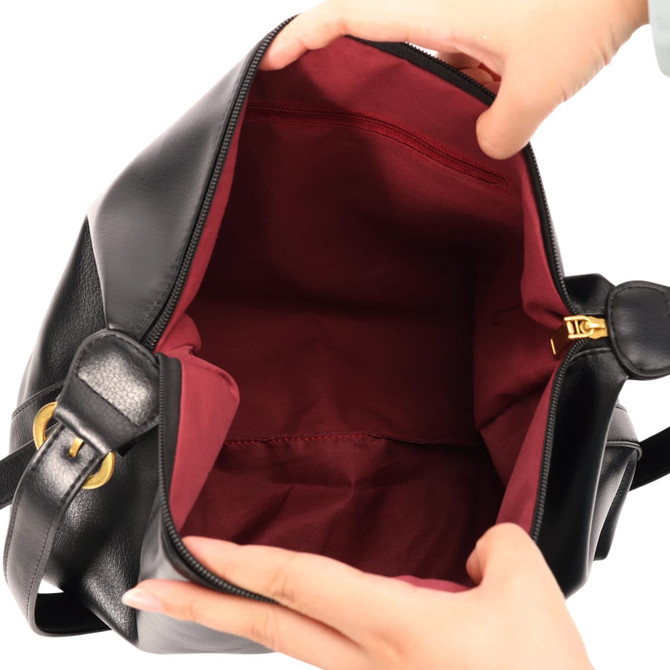 A black tote bag opened to reveal a burgundy lining, held by a pair of hands. The bag features a gold zip closure.