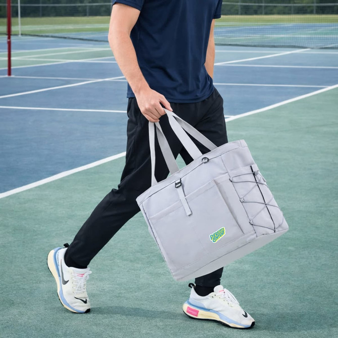 A grey lightweight tennis tote bag with handles, featuring a logo and elastic bungee cord, carried by a person on a tennis court.