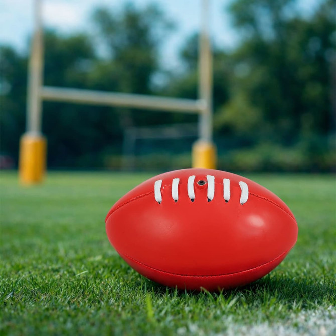 A red mini promotional Aussie Rules ball resting on green grass, with goalposts blurred in the background.