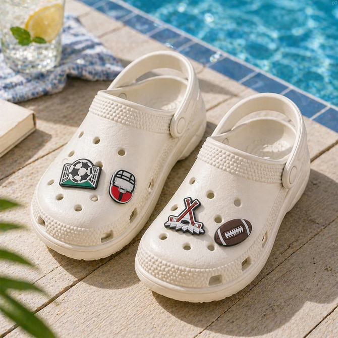 A pair of white Crocs with four custom shoe charms featuring sports designs, set near a poolside.