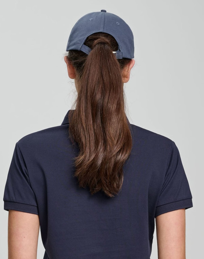 A woman with long hair wears a navy blue unstructured cap, viewed from the back. The cap features a branding logo.