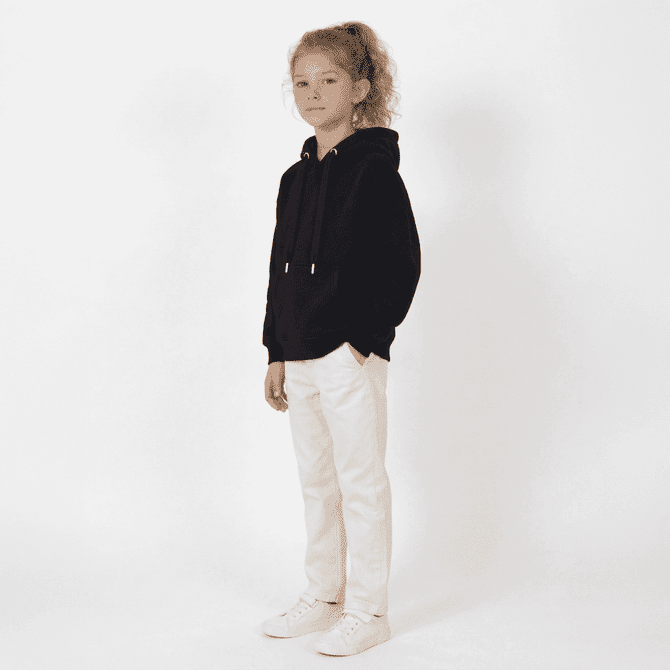 A young girl stands wearing a black hooded sweatshirt and white pants, with a light background. The hoodie has a logo.