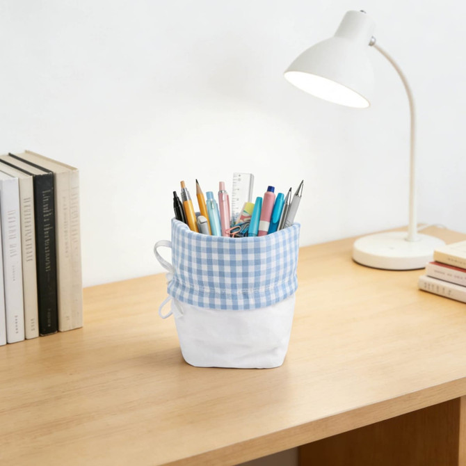 A foldable drawstring pencil case in blue and white gingham, filled with various pens and stationery on a wooden desk.