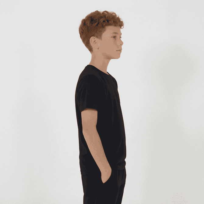 A black youth tee is displayed on a young person standing sideways with hands in pockets.
