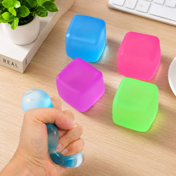 A hand squeezing a blue squishy stress relief cube among pink, green, and blue cubes on a wooden desk.