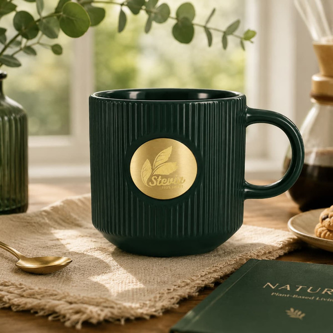 A green ceramic mug with a ribbed design and a gold medal-like logo sits on a textured surface, alongside a spoon and snacks.
