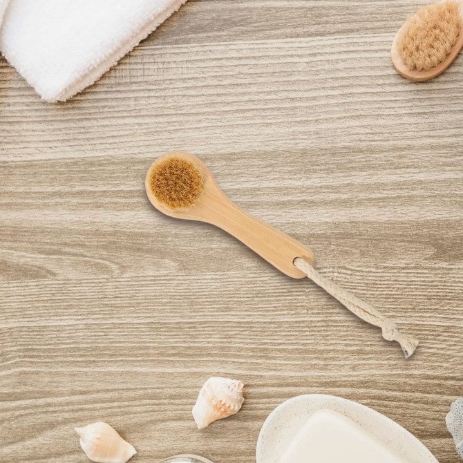 A natural wooden face brush with a beige bristle head, set against a wooden background with soft towels and seashells.