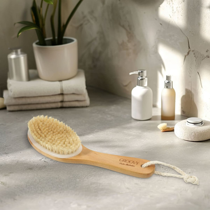 A dry body brush with a wooden handle and bristles on a countertop, surrounded by skincare products and towels.