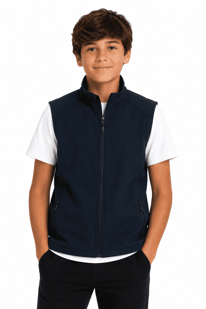 A boy stands wearing a navy blue vest over a white T-shirt, with his hands in his pockets. The vest has branding.